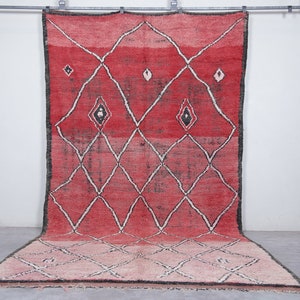 Op de afbeelding: Een rood en roze Marokkaans tapijt met een ruitpatroon en zwarte accenten. Het tapijt hangt aan een muur en heeft een franje rand.