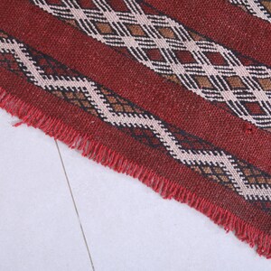 May include: Close-up of a red, brown, black, and white woven rug with a fringe border. The rug has a geometric pattern.
