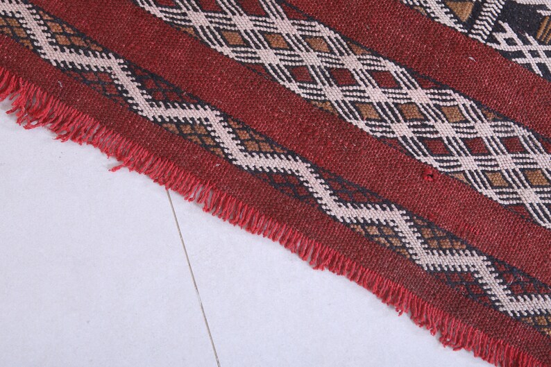 May include: Close-up of a red, brown, black, and white woven rug with a fringe border. The rug has a geometric pattern.