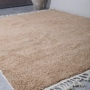 May include: A light brown shag rug with a white fringe border. The rug is on a gray tiled floor.