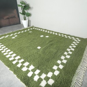 May include: A green rectangular rug with a white checkered border and fringe. The rug has a soft, textured appearance and is placed on a gray floor. The rug's dimensions are not visible in the image.