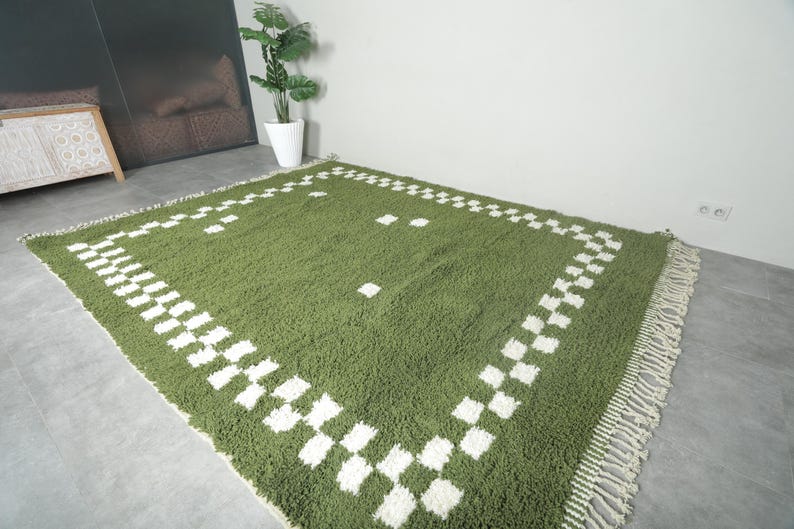 May include: A green rectangular rug with a white checkered border and fringe. The rug has a soft, textured appearance and is placed on a gray floor. The rug's dimensions are not visible in the image.