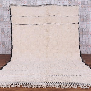May include: A white wool rug with black trim and fringe. The rug has a woven pattern and is made in Morocco.