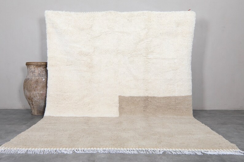 Puede incluir: Una alfombra de pelo blanco y beige con borde de flecos. La alfombra tiene un dise&ntilde;o bicolor con una secci&oacute;n superior blanca y una secci&oacute;n inferior beige.