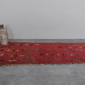 May include: A red, hand-woven rug with a geometric pattern in black and yellow. The rug has a fringe border and is laid out on a gray floor.