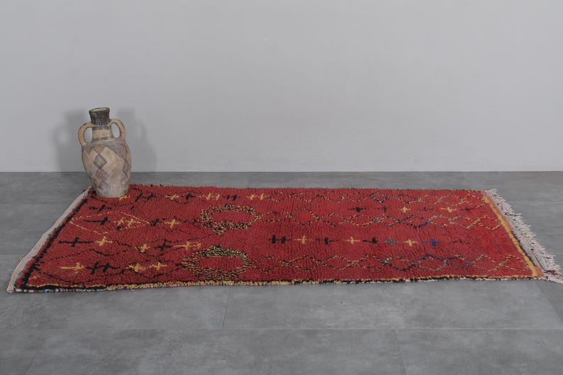 May include: A red, hand-woven rug with a geometric pattern in black and yellow. The rug has a fringe border and is laid out on a gray floor.