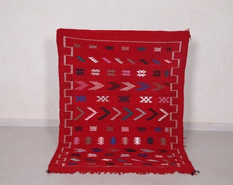 Op de afbeelding: Een rood Marokkaans tapijt met een geometrisch patroon in tinten blauw, groen, roze en wit. Het tapijt heeft een franje rand.
