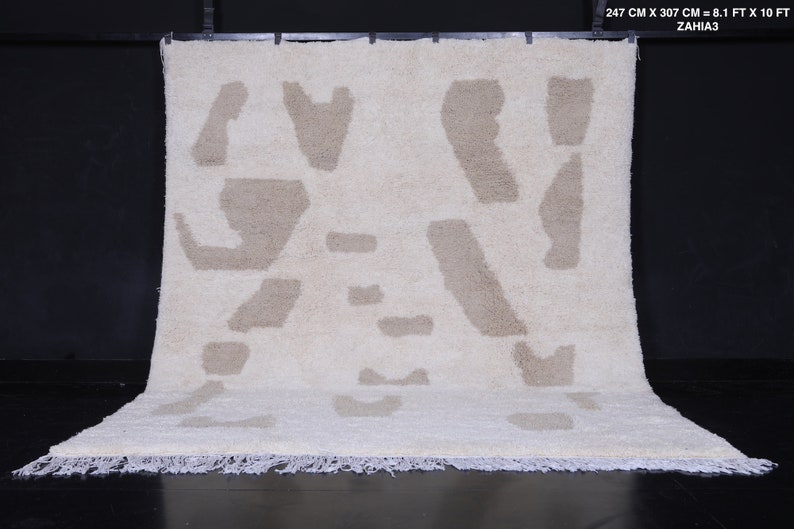 May include: A white rug with a beige abstract pattern. The rug has a fringe border and measures 8.1 feet by 10 feet.