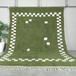 May include: Olive green rug with a white checkered border and scattered white squares. The rug has a shaggy texture and a fringe along the bottom edge. A potted plant is in the background.