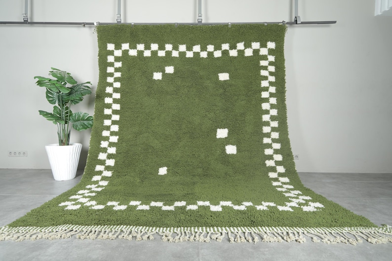 May include: Olive green rug with a white checkered border and scattered white squares. The rug has a shaggy texture and a fringe along the bottom edge. A potted plant is in the background.