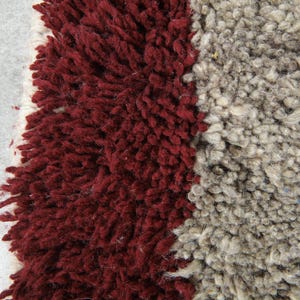 May include: Close-up of a textured rug with a mix of colors. The rug features a deep burgundy section on the left, a light gray section on the right, and a white border. The rug's texture is shaggy and plush.