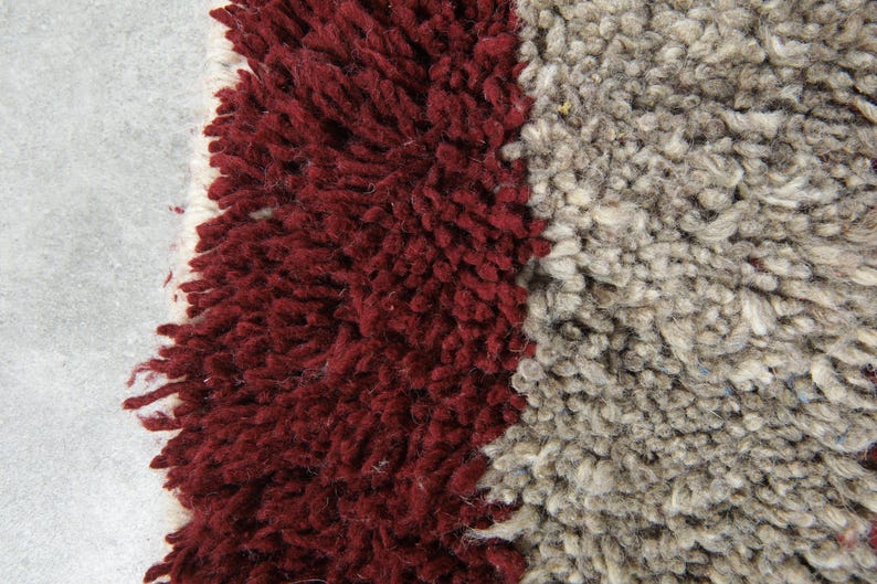 May include: Close-up of a textured rug with a mix of colors. The rug features a deep burgundy section on the left, a light gray section on the right, and a white border. The rug's texture is shaggy and plush.