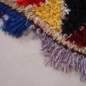 May include: Close-up of a colorful woven rug with fringe. The rug is made of different colored yarns, including red, yellow, black, and white. The fringe is a mix of blue, purple, and white.