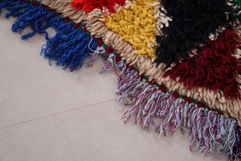 May include: Close-up of a colorful woven rug with fringe. The rug is made of different colored yarns, including red, yellow, black, and white. The fringe is a mix of blue, purple, and white.