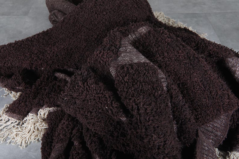 May include: A dark brown, textured throw blanket with a shaggy appearance. The blanket has a woven backing and a cream-colored fringe along the edges. The fabric appears soft and plush.