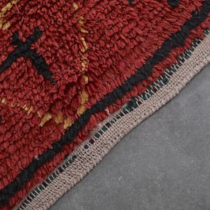 May include: Close-up of a red, black, and gold patterned rug with a fringe border. The rug is made of a thick, textured material.