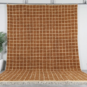 Puede incluir: Una alfombra grande y rectangular con un patrón de cuadrícula en tonos marrones y crema. La alfombra tiene un borde de flecos.