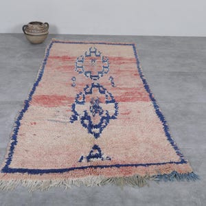 May include: A vintage Moroccan rug with a cream background and a blue and pink geometric pattern. The rug has a fringe border and is made of wool.