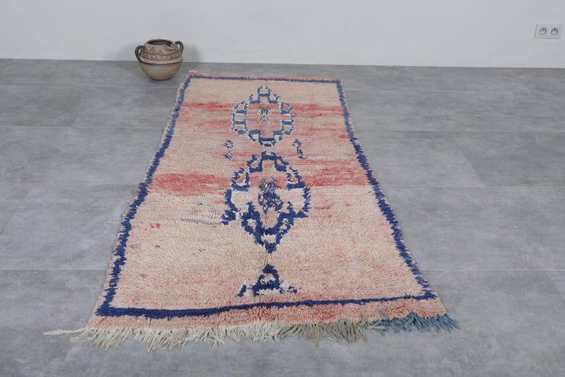May include: A vintage Moroccan rug with a cream background and a blue and pink geometric pattern. The rug has a fringe border and is made of wool.