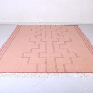 May include: A light pink rug with a geometric pattern woven into the fabric. The rug has a white fringe around the edges.