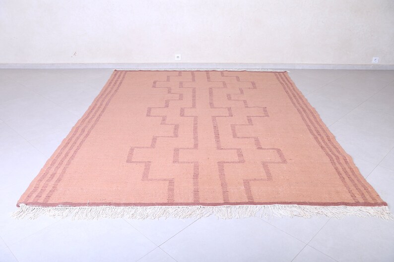May include: A light pink rug with a geometric pattern woven into the fabric. The rug has a white fringe around the edges.