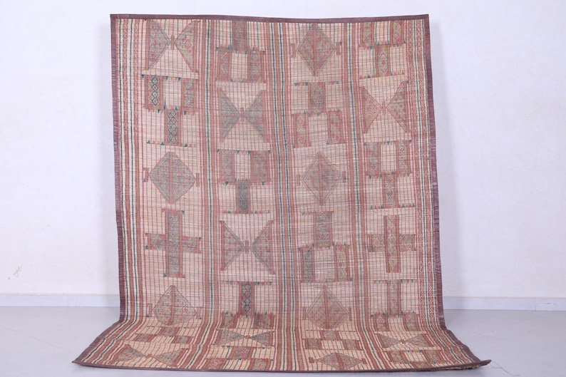 May include: A rectangular woven mat with a geometric pattern in shades of beige, red, and brown. The design features repeating shapes including crosses, diamonds, and triangles. The mat has a dark brown border.