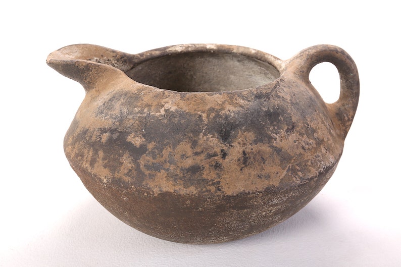May include: A brown, unglazed, earthenware pitcher with a rounded body and a single handle. The pitcher has a wide, open mouth and a slightly flared lip.