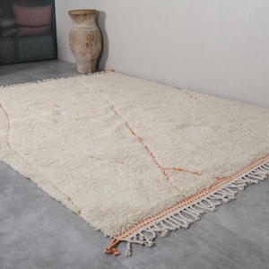 May include: A large, rectangular, off-white rug with a shaggy texture. The rug features a simple design with thin, orange lines and a fringed border. The rug is placed on a gray floor, and the overall aesthetic is neutral and inviting.