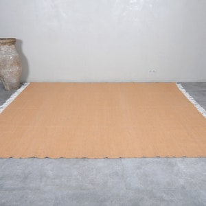 May include: A light brown rectangular rug with a white fringe border. The rug is laid out on a gray tiled floor.