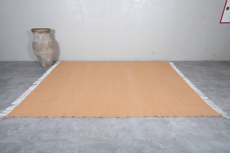 May include: A light brown rectangular rug with a white fringe border. The rug is laid out on a gray tiled floor.