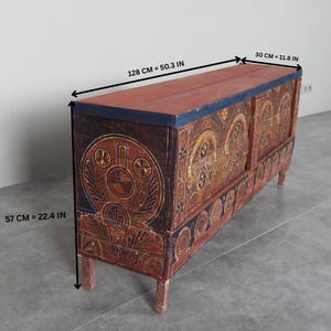 May include: An antique wooden chest with a red top and ornate, hand-painted designs. The chest is 128 cm long, 30 cm wide, and 57 cm high. The chest has a dark blue trim and is supported by four short legs.