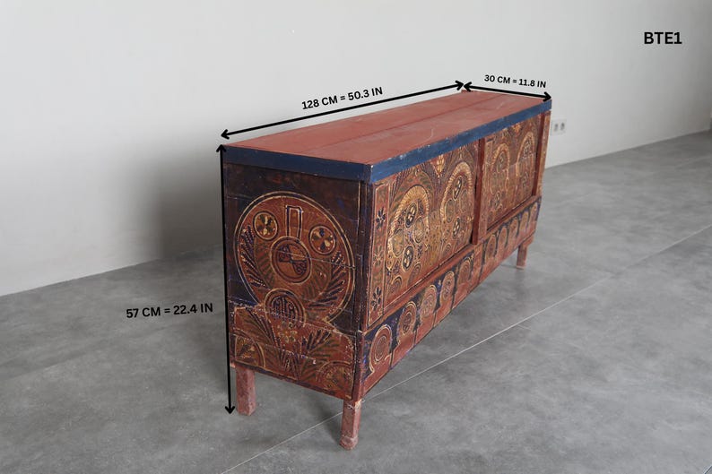 May include: An antique wooden chest with a red top and ornate, hand-painted designs. The chest is 128 cm long, 30 cm wide, and 57 cm high. The chest has a dark blue trim and is supported by four short legs.
