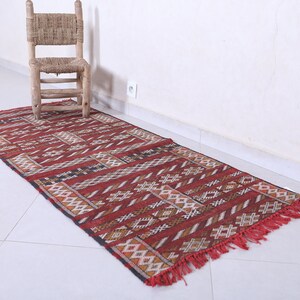 May include: A red, black, and white patterned runner rug with fringe. The rug is laid out on a white tiled floor.