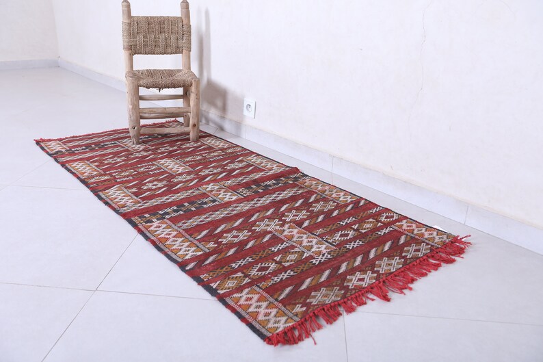 May include: A red, black, and white patterned runner rug with fringe. The rug is laid out on a white tiled floor.