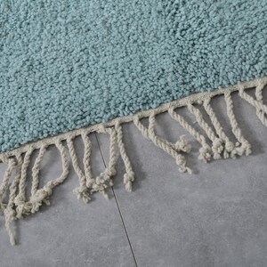 May include: A light blue rug with a fringe border. The fringe is made of white cotton rope.