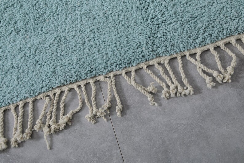 May include: A light blue rug with a fringe border. The fringe is made of white cotton rope.