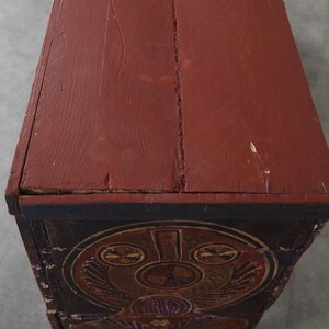 May include: A close-up of a wooden chest with a red-brown painted top. The chest features decorative carvings and a dark blue border. The top shows signs of wear and age, with visible cracks and imperfections.