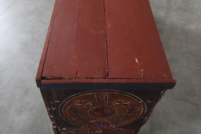 May include: A close-up of a wooden chest with a red-brown painted top. The chest features decorative carvings and a dark blue border. The top shows signs of wear and age, with visible cracks and imperfections.