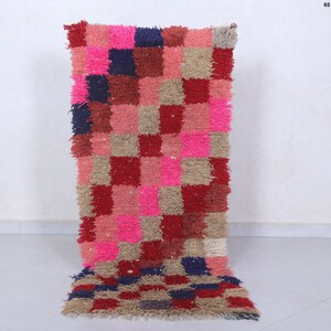 May include: A long, rectangular rug with a checkered pattern in shades of pink, red, beige, and blue. The rug is made of a fluffy, textured material.
