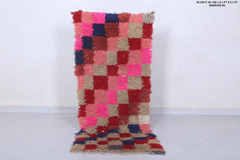 May include: A long, rectangular rug with a checkered pattern in shades of pink, red, beige, and blue. The rug is made of a fluffy, textured material.