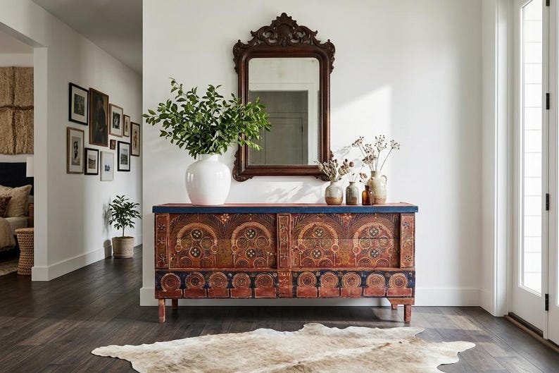 May include: A detailed wooden chest with ornate carvings and a distressed finish. A white vase with greenery sits on top, with a dark brown framed mirror above. The scene is completed with a rug and other decorative items.