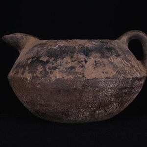 May include: A brown earthenware jug with a rounded body and a single handle. The jug has a spout on one side and is slightly weathered.