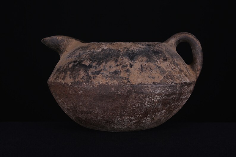 May include: A brown earthenware jug with a rounded body and a single handle. The jug has a spout on one side and is slightly weathered.