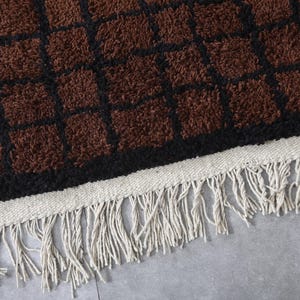 Peut inclure: Gros plan sur un tapis &agrave; motifs marron et noir avec une surface textur&eacute;e. Le tapis pr&eacute;sente un motif quadrill&eacute; avec des carr&eacute;s marron soulign&eacute;s de noir. Le bord a une bordure tiss&eacute;e blanche avec de longues franges cr&egrave;me.