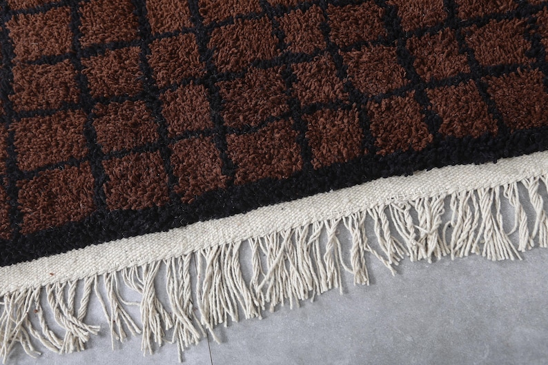 Peut inclure: Gros plan sur un tapis &agrave; motifs marron et noir avec une surface textur&eacute;e. Le tapis pr&eacute;sente un motif quadrill&eacute; avec des carr&eacute;s marron soulign&eacute;s de noir. Le bord a une bordure tiss&eacute;e blanche avec de longues franges cr&egrave;me.