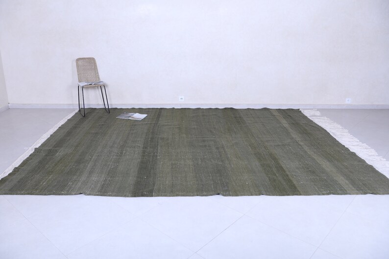May include: A green woven rug with a white fringe border. The rug is laid out on a white tiled floor.