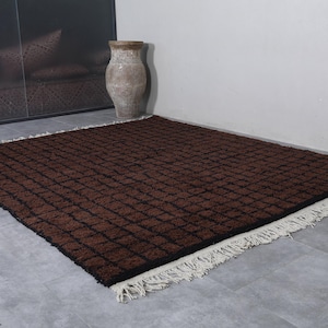 Peut inclure: Un tapis rectangulaire marron avec un motif quadrill&eacute; et des franges blanches. Le tapis est pos&eacute; sur un sol gris, avec un grand vase et un mur marron fonc&eacute; en arri&egrave;re-plan. Les dimensions du tapis ne sont pas visibles.