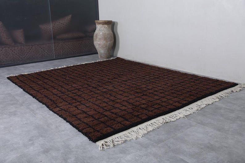 Peut inclure: Un tapis rectangulaire marron avec un motif quadrill&eacute; et des franges blanches. Le tapis est pos&eacute; sur un sol gris, avec un grand vase et un mur marron fonc&eacute; en arri&egrave;re-plan. Les dimensions du tapis ne sont pas visibles.