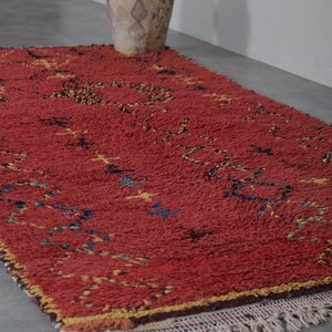 May include: A red Moroccan rug with a geometric pattern in yellow, blue, and black. The rug has a fringe border and is lying on a gray floor.