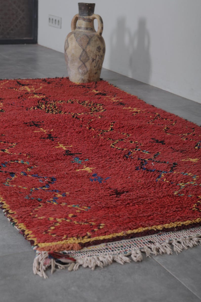 May include: A red Moroccan rug with a geometric pattern in yellow, blue, and black. The rug has a fringe border and is lying on a gray floor.